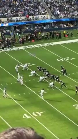 The video shows a football game from an elevated perspective. The teams are lined up in formation on a green field marked with yard lines. Players are wearing uniforms; one team is in white and the other in dark colors. The play begins, and the players engage in blocking and tackling. The environment is a packed stadium with many spectators in the stands. The atmosphere is energetic.