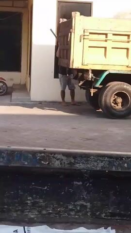A man wearing a black t-shirt and patterned shorts is standing next to the back of a yellow dump truck. He is holding a long, dark pole. He is using the pole to lift the rear door of the truck. The man then walks away from the truck, carrying the pole over his shoulder. In the background, there is a building with a white wall and a red motorcycle parked near it. The ground is paved with concrete.