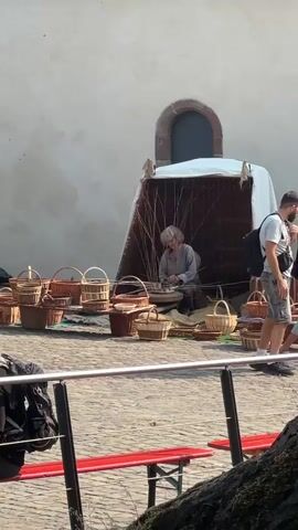 A person is sitting inside a woven structure, weaving a basket. The person has light-colored hair and is wearing a grey shirt. Various woven baskets are displayed in front of the structure. Several people are walking around the area, some of whom are looking at the basket weaver. The ground is paved with cobblestones, and a light-colored building with a dark arched doorway is visible in the background. Red benches and a metal railing are also present in the foreground.