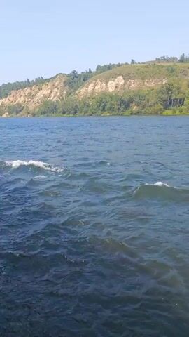 A slightly overcast day with calm blue water with light waves rippling across it. A lush green hillside with trees covers the background, meeting a clear blue sky at the top. The video pans slightly to the right, revealing more of the water and hillside. There are no people or objects visible in the video.