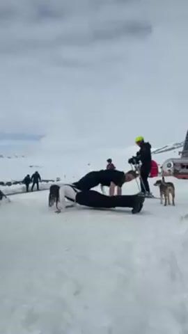 In a snowy outdoor setting with mountains visible in the background, a man and a woman are performing tandem push-ups. The woman, wearing a white top and dark pants, is positioned on top of the man, who is wearing all black. Several other people, dressed in ski attire, are standing or walking around them. Ski equipment is scattered nearby. A small, light-brown dog is also present, standing to the side and watching. The sky is overcast.