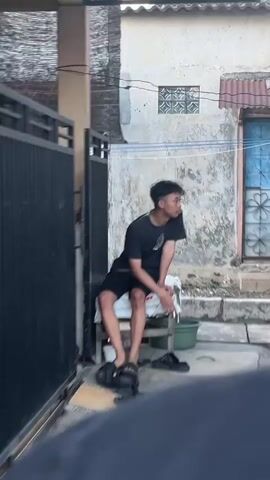 A young man wearing a black t-shirt and black shorts is sitting on a white plastic chair in front of a textured wall. He has black hair and is looking to his right. He is wearing black sandals. To his left, a dark metal gate is visible. A green bucket is on the ground near his feet. The background shows a building with a tiled roof and a barred window. The lighting suggests an outdoor environment during the daytime.