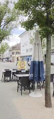The video shows a street scene in a town or city. There are buildings lining the street, some of which appear to be shops or restaurants with outdoor seating areas. Several tables and chairs are set up on a paved sidewalk. Trees line the street, providing shade. A tall lamppost is visible in the foreground. People are walking along the sidewalk and crossing the street in the distance. Cars are parked along the street and driving in the background. The sky is visible, with some clouds. The overall impression is of a moderately busy, European-style street during the daytime.