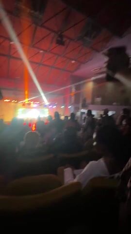 The video shows an indoor event, likely a concert or party, with a large crowd of people. The setting appears to be a hall or auditorium with a high ceiling featuring a metal truss structure. Bright stage lights in orange, white, blue, and green illuminate the stage area where several individuals are visible. The crowd is actively engaged, with many people standing, dancing, and clapping to the music. Some attendees are seated in the foreground, and one person can be seen holding a phone, possibly recording the event. The atmosphere is energetic and lively, suggesting an evening or nighttime event. No visible text is present in the video. The event is taking place in Ife, Nigeria.