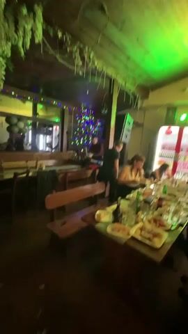 The video shows people enjoying themselves at a restaurant. The room is decorated with twinkling lights and fake foliage. Some people are seated at tables, eating and talking. Others are standing and dancing to the music. The lighting changes colors throughout the video, adding to the festive atmosphere.