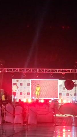 The video shows a runway with a woman in a gold outfit standing in the center, posing for a photo. The runway is lit with red lights and a spotlight. There are several people surrounding the stage, including a man standing on the left side and two men standing on the right side of the stage. There is also a large screen behind the woman with a red wavy design on it. The video is slightly shaky and blurry. The scene appears to be from a fashion show or similar event.