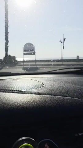 The video is captured from inside a car looking through the windshield and passenger window. The sun is bright and shining, with some glare on the windshield. Outside the car, there are palm trees, streetlights, and a body of water, likely the ocean, in the distance. The car appears to be moving. The interior of the car, including the dashboard and a side mirror, is visible. The overall impression is a sunny day drive along a coastal road.