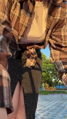 A young woman stands outside in an open area with tiled ground, likely a park or playground, with a blue sky overhead. She is wearing a long black denim skirt with a front slit, a white t-shirt, and a brown plaid shirt tied at the waist. She has short brown hair and is wearing a necklace. Initially, she faces the camera, adjusts her hair, and then turns her back, placing her hands on her hips. In the background, there are trees and playground equipment. The lighting suggests it is either early morning or late afternoon due to the sun's angle.