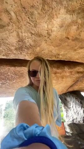 A person with long blonde hair and sunglasses is standing in front of a waterfall, holding a camera to take a selfie. The person is wearing a light blue t-shirt. The waterfall is surrounded by rocks and vegetation. The scene appears to be a natural setting, possibly a park or a tourist attraction. The lighting is bright, suggesting it is daytime.