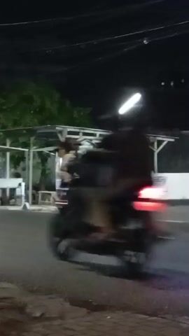 A person in a red shirt and gray pants is walking near a food stall. A motorcycle drives past, with two people on it, one of them blurry. The scene is at night, with lights illuminating the food stall.