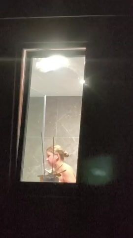The video shows a person from the shoulders up standing in a shower stall. The person is facing to the left and has blonde hair pulled back in a bun. The person is holding a light blue object, possibly a loofah or washcloth, and is scrubbing their upper body. The shower stall has marble-patterned tiles on the wall and a glass door. The room is dimly lit with a bright light visible above the shower. The video appears to be taken from outside the shower area, looking through a window.