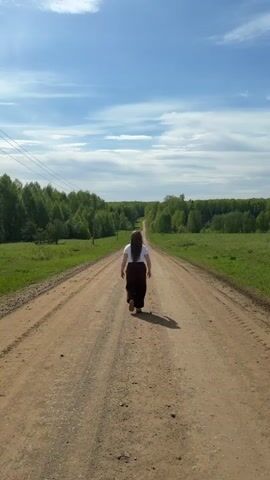 A woman with long dark hair is seen from behind, walking down a long, dirt road. She is wearing a white t-shirt and dark pants. The road stretches into the distance, with fields and trees on either side. The sky is blue with some clouds. At one point, the woman spreads her arms out to the sides as she walks. There are power lines on the left side of the road.