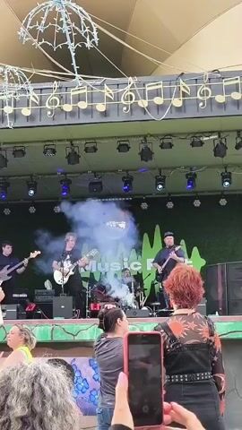 A band is performing on a stage. There are four musicians on stage: a bassist on the left, a guitarist in the center playing a white guitar, a drummer behind them, and another guitarist on the right playing a black guitar. There is smoke on the stage, and a disco ball is visible. The stage has a backdrop with "Music Fest" written on it in large green letters. The stage is decorated with green foliage along the bottom and has lights above it. There are also two umbrella-shaped light fixtures hanging from the ceiling structure above the stage. In front of the stage, a crowd of people are watching the performance. Two people in the foreground are holding up their phones to record the concert. One person is holding a large white sign.