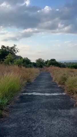 A paved path winds uphill through dry grass and trees. The sky above is partly cloudy. The video is shot from a first-person perspective.