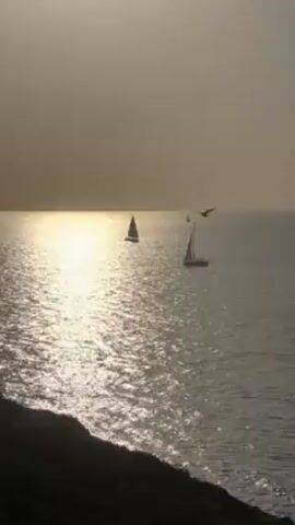 The video shows a seascape at what appears to be dusk or dawn, with the sun low on the horizon, creating a bright reflection on the water. Two sailboats are visible in the distance, each with its sails up, suggesting they are underway.  A third, smaller sail may be visible further to the right and away from the other two.  The foreground shows a dark, silhouetted hillside or cliff. The overall scene is serene and somewhat monochromatic due to the lighting conditions.  The water is choppy, with visible wavelets.