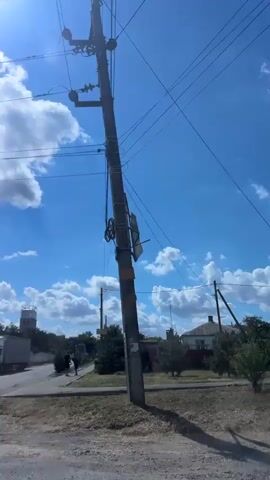 This video shows a view from a moving vehicle looking towards a landscape under a bright, sunny sky with scattered white clouds. In the distance, there is a tall, white industrial-looking structure, possibly a water tower or silo, with a smaller, smokestack-like structure next to it. Several utility poles with wires crisscross the frame. To the right of the industrial structures, there are houses and trees. Closer to the foreground, there is a roadside with dry grass and a paved surface. A small tree is visible in the middle ground. A sign with Cyrillic lettering, possibly indicating a location or business, is visible on a building to the right. A white truck with a cargo container is parked on the left side of the frame. Two figures are standing near the truck. The overall scene suggests a rural or semi-industrial area.