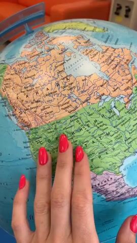 A hand with bright red fingernails hovers over a globe, appearing to point at various locations. The globe is detailed, showing continents, countries, cities, and bodies of water. The background is blurred, but suggests an indoor setting with an orange piece of furniture visible. The video cuts between views of North America, the Atlantic Ocean, Europe, and part of Africa.