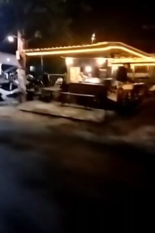 The video shows a street scene at night. Several motorcycles are parked along the side of the road, some in front of food stalls or small shops that are illuminated by string lights. People can be seen walking and standing around. The camera is moving, suggesting the video is being recorded from a vehicle or while someone is walking. The lighting is dim, with the primary light sources coming from the shops and streetlights. The ground appears to be paved. There are trees and poles visible along the roadside.