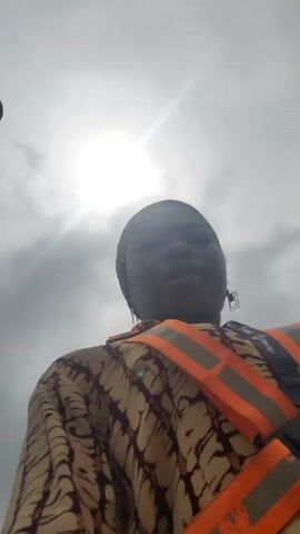 A woman wearing a headscarf, a patterned shirt, and a high-visibility vest is looking at the camera. She is outdoors, with the sun and clouds visible in the sky behind her. The woman is speaking to the camera, and the audio appears to be of her singing.