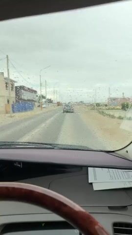 The video shows the view from inside a car on a rainy day. The windshield is covered in raindrops. The car is driving on a paved road. There are buildings and power lines visible on the side of the road. There are other cars on the road ahead. The steering wheel of the car is visible in the foreground. The video cuts out briefly as the windshield wipers activate.