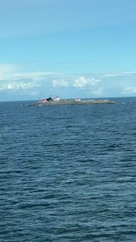 A drone shot slowly pans across a vast expanse of deep blue water. In the distance, a small, rocky island emerges from the sea, dotted with several small buildings with red roofs. A prominent lighthouse stands tall on the island, its white structure contrasting with the dark rocks and the surrounding ocean. The sky above is a clear, bright blue, with a few wispy white clouds scattered across the horizon. The overall scene is serene and picturesque, evoking a sense of isolation and natural beauty.
