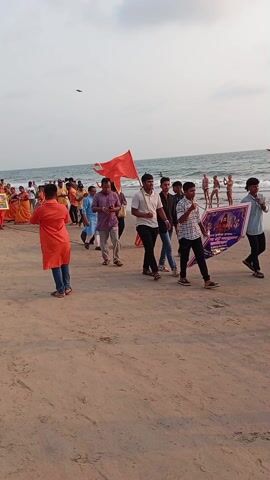 A group of people are walking along a beach. Most of the people are wearing orange clothing or carrying orange flags. There are also a few children running around in the crowd. Some of the people are carrying a framed picture and a banner with images on it. The beach is sandy, and the ocean is visible in the background. The sky is overcast.