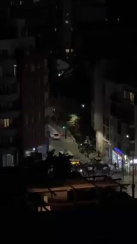 Here's a description of the video:

The video shows a nighttime view of a city street from an elevated position. The street is lined with buildings, some of which have illuminated windows. Streetlights provide additional illumination. Cars are seen driving on the street, with their headlights visible. There are also trees and foliage visible along the street. The overall scene is a typical urban environment at night.