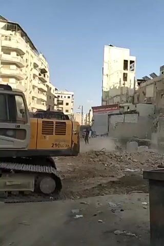 The video shows a yellow Hyundai 210LC-7A excavator operating in a debris-strewn urban environment. The excavator is positioned amidst rubble and damaged structures, with taller buildings visible in the background. The operator is seen inside the excavator's cabin, manipulating the controls. The excavator's arm and bucket are in motion, interacting with the debris on the ground. A white car is seen in the background, and the minaret of a mosque is visible rising above the surrounding buildings.