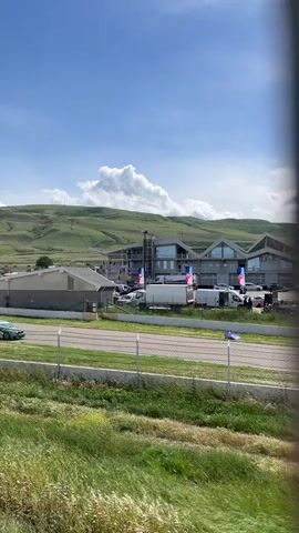 The video shows a race track, likely during a race or practice session. Several race cars are seen speeding along the track. In the background is a complex of buildings that might be related to the track facilities, with what appear to be flags displayed around the area. Behind the buildings is a green, hilly landscape under a blue sky with white clouds. The camera appears to be behind a fence and some vegetation. The weather seems sunny.