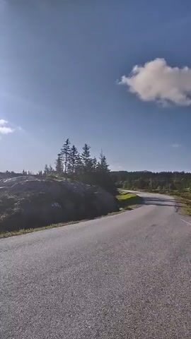 The video shows a road with a rocky, tree-covered landscape on the left. The sky is blue with scattered clouds, and the sun is bright. The camera is moving forward, suggesting it is attached to a vehicle or being held by someone walking. The road appears to be paved and curves to the right. The landscape is natural, with trees and grass, and the overall impression is one of a scenic, possibly rural, environment. There are no people or animals visible in the video.