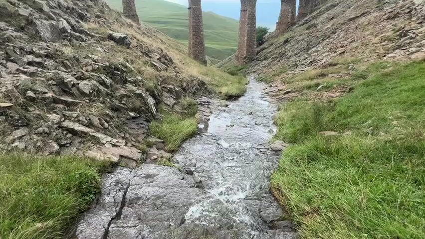 A narrow, rocky stream flows through a steep, grassy ravine. On the left, a rocky slope descends towards the stream. On the right, a similar slope rises, covered in grass. In the distance, a curved, stone viaduct with multiple arches spans the ravine. The sky is overcast. The camera is positioned at the stream level, looking upwards towards the viaduct. The video is a continuous, static shot from a single camera angle.