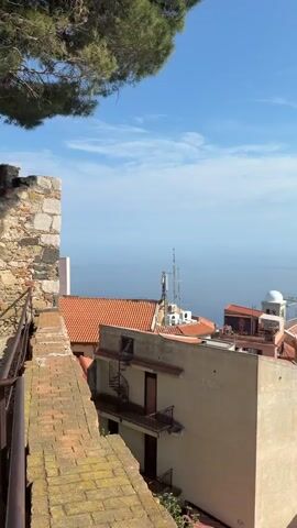 The video shows a panorama of a coastal area with buildings, hills, and a mountain range in the distance.  The foreground is dominated by rooftops of buildings with orange clay tiles. There is a view of the coastline and the sea, which stretches out to the horizon under a bright blue sky with some scattered clouds. The camera then pans to reveal a series of hills and mountains, covered in green vegetation. In the background, a snow-capped mountain range is partially obscured by clouds. The weather appears sunny and clear overall.