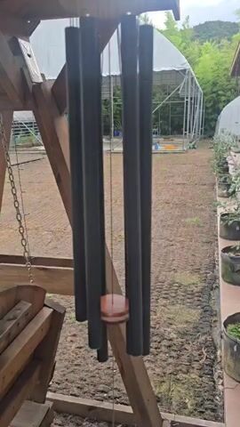 A close-up shot focuses on a set of dark metal wind chimes hanging from a wooden structure. The chimes have a circular wooden piece at the top and a single circular wooden clapper in the middle. The background shows a muddy ground, a greenhouse with a white, arched roof, and some green foliage. A wooden swing set is partially visible on the left side of the frame. The video appears to be a still shot with no observable movement.