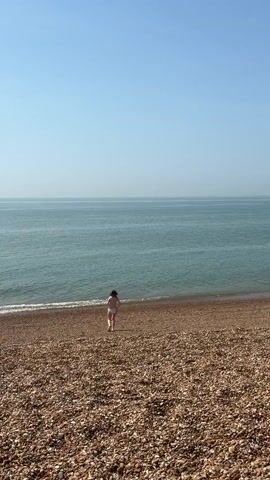 The video shows a young woman walking towards the ocean on a pebble beach. The woman has dark hair and is wearing a pink bikini. The ocean is calm and turquoise in color, with small waves lapping at the shore. The sky is clear and blue. The beach is composed of small, rounded pebbles in shades of brown and tan.