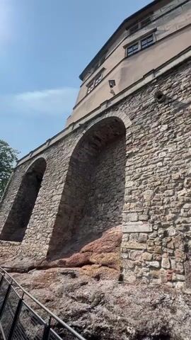 The video pans up a stone wall towards a building. The wall is made of rough, uneven stones of various sizes and colors, with some reddish-brown rock formations at the base. There are three large arched openings in the wall, recessed and filled with more stone. Above the stone wall is a lighter-colored building with several windows. The sky is blue with a few wispy clouds. A black railing is visible in the bottom left corner of the frame.