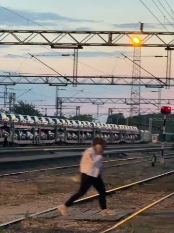 A person walks across railroad tracks while talking on a cell phone. The person is wearing a light-colored jacket and dark pants. A train carrying cars is visible in the background. Overhead are metal structures supporting power lines. A signal light is red. The sky is a mix of colors suggesting dusk or dawn.