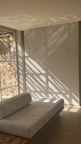 The media file depicts a minimalist indoor scene bathed in warm, natural light. A white daybed with a cylindrical bolster pillow dominates the foreground, positioned on a light-colored wooden platform. The daybed is oriented towards a large window with a stone wall visible on the other side. The wall directly opposite the window has a textured, speckled finish and is adorned with dramatic, dappled shadows, suggesting the presence of foliage outside filtering the sunlight.

The lighting indicates a sunny day, likely in the late morning or early afternoon, given the strong diagonal cast of the shadows. The overall ambiance is serene and tranquil, evoking a sense of peace and relaxation. The setting appears to be a modern, uncluttered living space or lounge area, possibly within a vacation villa or a spa, given the natural materials like stone and wood, and the emphasis on light and comfort. There is no visible text in the media file. The absence of people and any specific action further contributes to the calm and introspective mood. The location context of Artà, Spain, suggests a Mediterranean environment, which aligns with the visual cues of sunlight, stone, and possibly lush vegetation casting shadows.