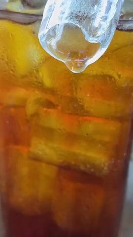 A close-up shot shows a plastic cup filled with iced tea. Large ice cubes occupy the upper portion of the cup, and condensation is visible on the exterior surface of the glass. The tea itself is a deep amber color, darkening towards the bottom of the cup.  A large ice cube at the top of the cup is melting, with a single water droplet forming at the bottom. The background is slightly blurred, suggesting a shallow depth of field.