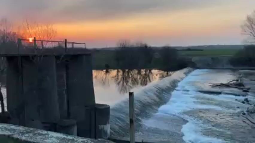 A static shot shows a dam at sunset.  The sun is barely visible, hidden behind trees in the background. The water in the dam is still enough to reflect the trees and sky. Water rushes over the dam, churning into white rapids. Logs are visible below the dam. The landscape beyond the dam is flat and green, with some trees.  The concrete dam structure is in the foreground to the left.