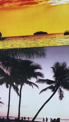 The video begins with a close-up of a framed picture depicting a tropical sunset over the ocean with palm trees silhouetted against the sky. The scene then transitions to an outdoor setting. A traditional wooden house with a tiered, pointed roof, characteristic of Southeast Asian architecture, is visible. The house is elevated on stilts. In the foreground, a dirt path winds through a yard with patches of grass and various plants. A light-colored pickup truck is parked near the house. Several wooden posts are positioned in the yard, some appearing to be part of an unfinished structure or decorative elements. A red and white traffic cone lies on the ground near a concrete trough that contains water. The sky is overcast, suggesting cloudy weather. The camera pans slightly, offering different perspectives of the house and its surroundings.