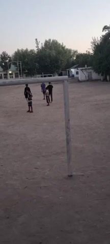 A group of children play soccer on a dusty field with a goalpost in the foreground. The sky is overcast, and the lighting suggests it is either early morning or late evening. The children are dressed in casual clothing. One child is kicking the ball, while others are observing or moving around the field. Trees and buildings are visible in the background, indicating an outdoor, possibly rural or suburban, setting. The ground is dirt and appears dry.