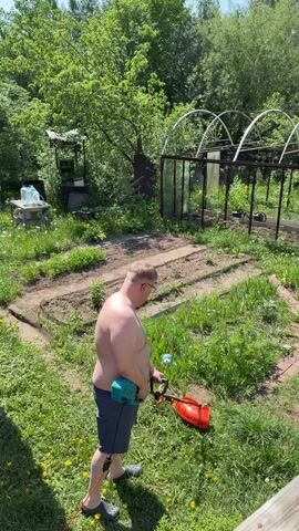 A shirtless man with blonde hair and glasses is mowing a lawn using a teal-colored electric grass trimmer. He is wearing grey shorts and black Crocs. The lawn area features garden beds, weeds, and dandelions. Behind the man is a greenhouse structure and some scattered gardening equipment. The camera pans to the right, revealing a wooden porch structure with a view of two tall, rusty metal silo-like structures, a small hut-like building covered with vegetation, and additional greenery. The sky is clear and blue.