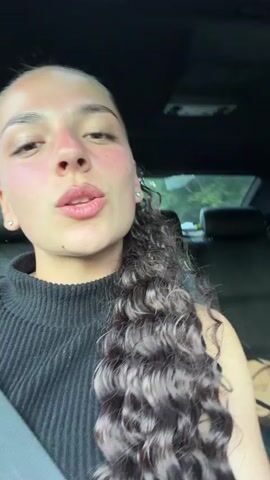 A young woman with curly dark hair and a black top films herself in a car. A man in a white t-shirt drives the car. The woman makes faces at the camera. The car appears to be moving through a wooded area, as evidenced by the blurred green foliage outside the windows.