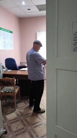 A man wearing a checkered shirt, black pants, and a cap stands in an office, holding a laptop and a piece of paper. He is looking down at the laptop. The office has a desk with papers, a chair, and an information board on the wall. A door is slightly ajar, framing the scene.