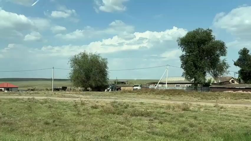 The video captures a view from a moving vehicle traveling along a dirt road through a rural area. The landscape is characterized by rolling hills covered in dry grass, with a few scattered trees and houses. Power lines run overhead, supported by poles. Several houses, some with red roofs and others with darker facades, are visible along the road. A blue tractor is parked near a large tree. In the foreground, the road is unpaved and appears to be a light brown color. The sky is blue with scattered white clouds, and sunlight illuminates the scene. A traffic sign depicting a quadruped animal is present on the left side of the road. The overall impression is one of a quiet, sparsely populated rural settlement under a bright sky.