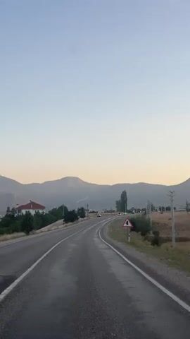 A vertical video shows a view from a vehicle traveling on a paved road. The road has white lines marking the lanes and appears wet. On the left side of the road, there are trees and a red-roofed house. In the background, there are mountains. On the right side of the road, there is a field, a tree, and a traffic sign indicating a sharp curve. The sky is clear with a light blue gradient. The video captures a sense of movement as the scenery shifts.