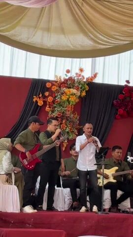 The video shows a band performing on a stage. The lead singer is a man wearing a white striped shirt and dark jeans, holding a microphone and singing into it. To his left is another man with glasses, playing a bass guitar. Behind him sits a drummer. To the singer's right is a man playing an electric guitar. They're all dressed in varying shades of green shirts. The stage has a burgundy colored backdrop, decorated with floral arrangements in orange, red, and green. The ceiling above is draped with white fabric.