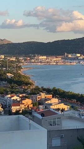This is a daytime, exterior, vertical shot of a coastal town and a bay. In the foreground, white and beige buildings with red-tiled roofs are visible, partially obscured by green trees. One building has a light-colored air conditioning unit attached to its exterior. The middle ground shows a bay with blue water, where several white sailboats are present. Along the edge of the bay, a shoreline with trees and a road is visible, with more red-tiled roof buildings. In the background, across the bay, is a large port or industrial area with numerous buildings, cranes, and docked ships. Behind the port, a large, tree-covered hill rises, and further in the distance, a mountainous landscape is visible. The sky is light blue with scattered white clouds, and the sun appears to be low in the sky, casting a warm light.