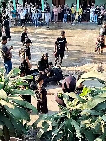 The video shows an outdoor scene with several people present, some of whom are wearing black clothing. In the foreground, a person in black clothing is lying on the ground and appears to be receiving attention from others also in black clothing. A young person in black clothing is standing and observing. In the background, a group of people are standing and watching, some of whom are seated. There are also some large green plants in the immediate foreground. Further in the background, there is a building and what appears to be a sound system. The ground is uneven and appears to be dirt or earth. The lighting suggests it is daytime. The overall impression is of an event or performance taking place.