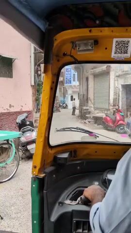 The video begins with a view from inside a yellow and green auto-rickshaw. The driver, a person with light-colored skin and wearing a light blue shirt and a dark baseball cap, is visible from the shoulders up, his right hand on the steering wheel. Through the windshield, a narrow street scene unfolds. Buildings with beige and pink walls line the street, some with closed metal shutters. A red scooter is parked on the left side of the street. Further down the street, two men are walking and a motorcycle is parked. One man wears a white garment, and another wears a dark helmet and jacket. The auto-rickshaw appears to be moving slowly through the street. A QR code is visible on the upper right side of the windshield. The video is shot from a first-person perspective, giving the viewer the sensation of being a passenger in the auto-rickshaw.